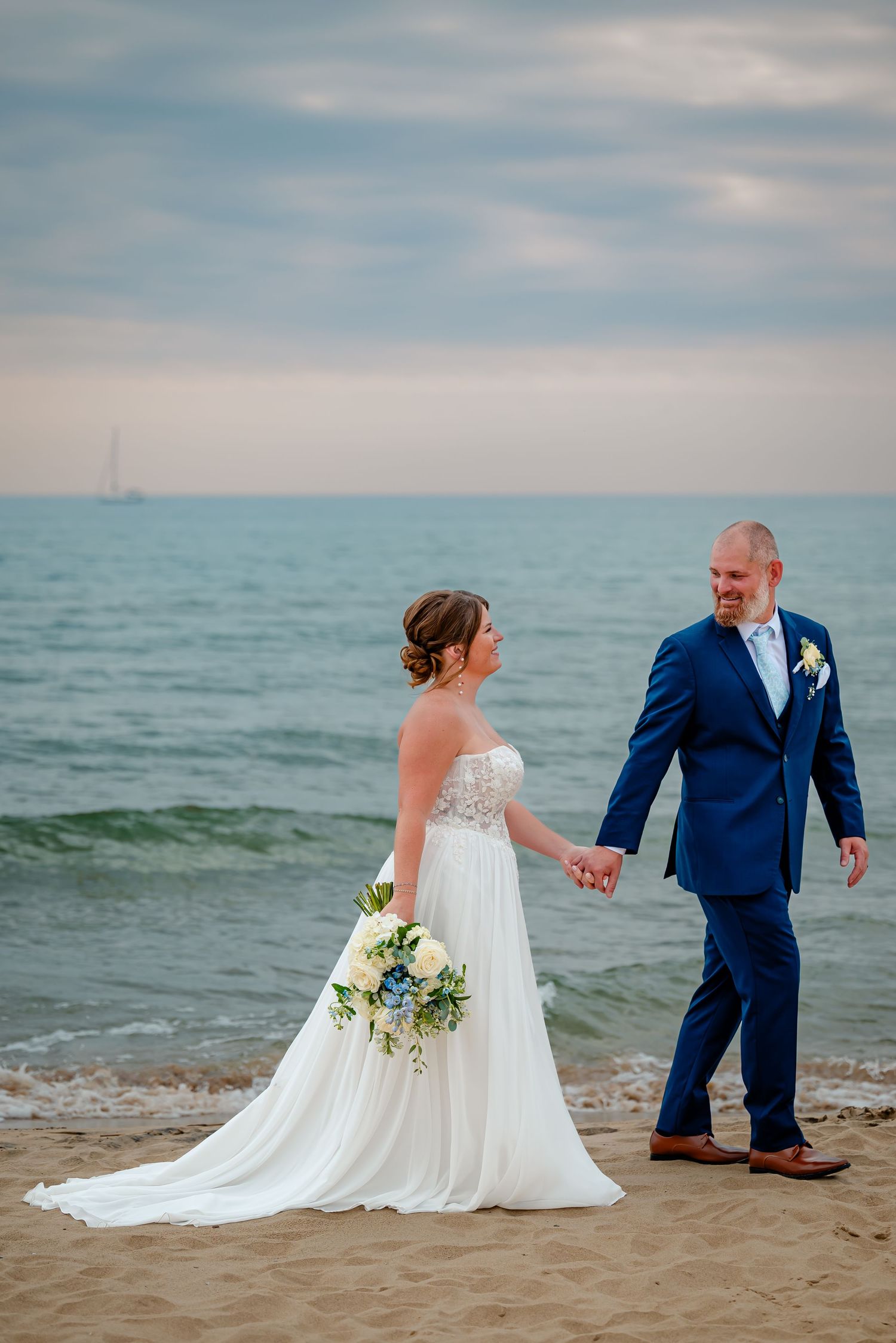 Newlyweds in elegant attire walk along sandy beach with ocean waves and cloudy sky in background.