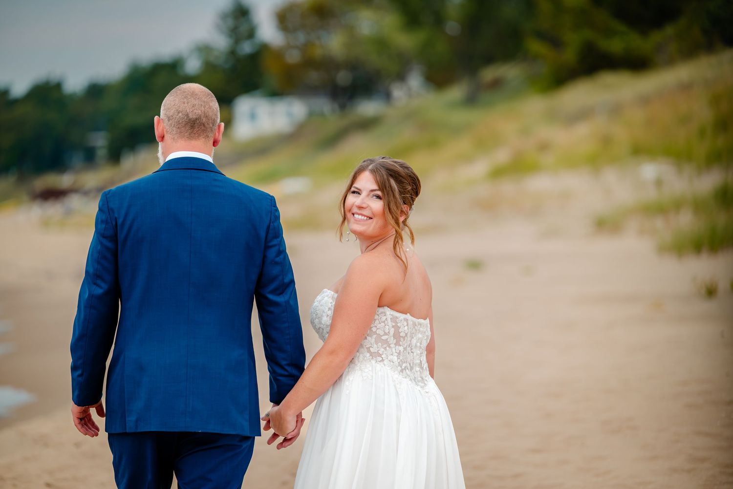 A couple walks together on a sandy beach path surrounded by grass and dunes on a sunny day.