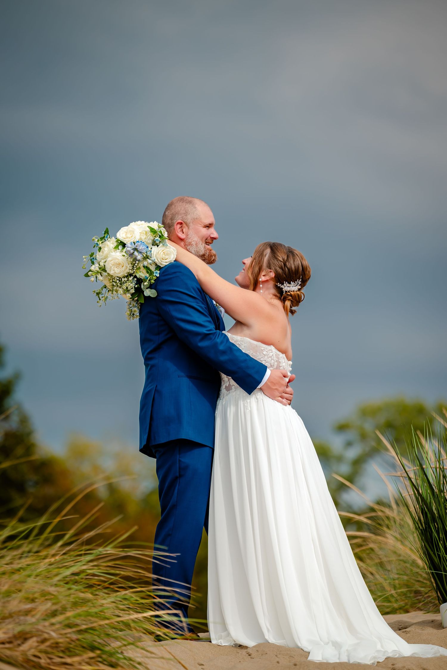 Newlywed couple embraces on a beach with tall grass swaying in the wind against a cloudy sky backdrop.