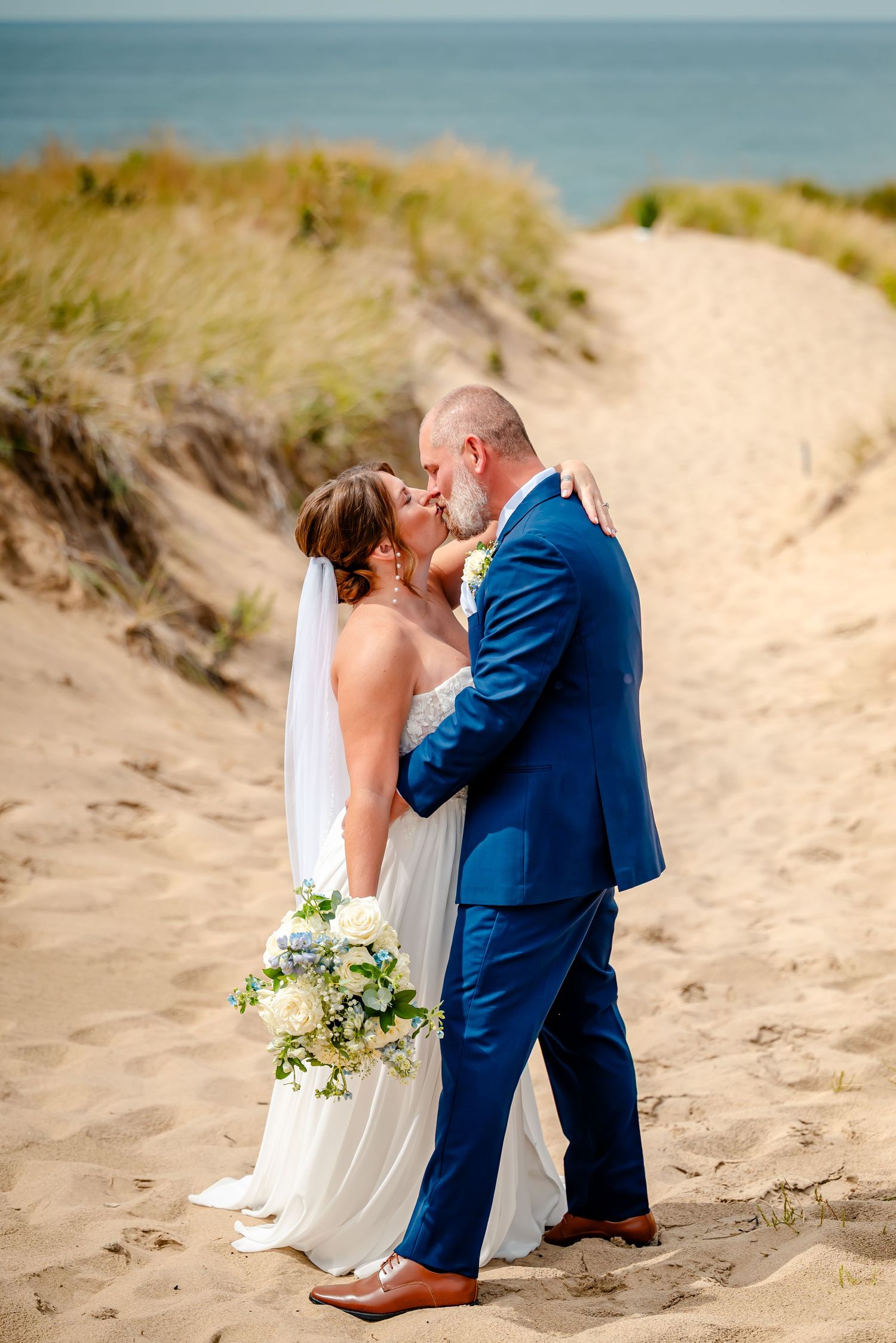 Romantic beach wedding couple embracing on sandy dunes with white bridal bouquet and navy blue suit.