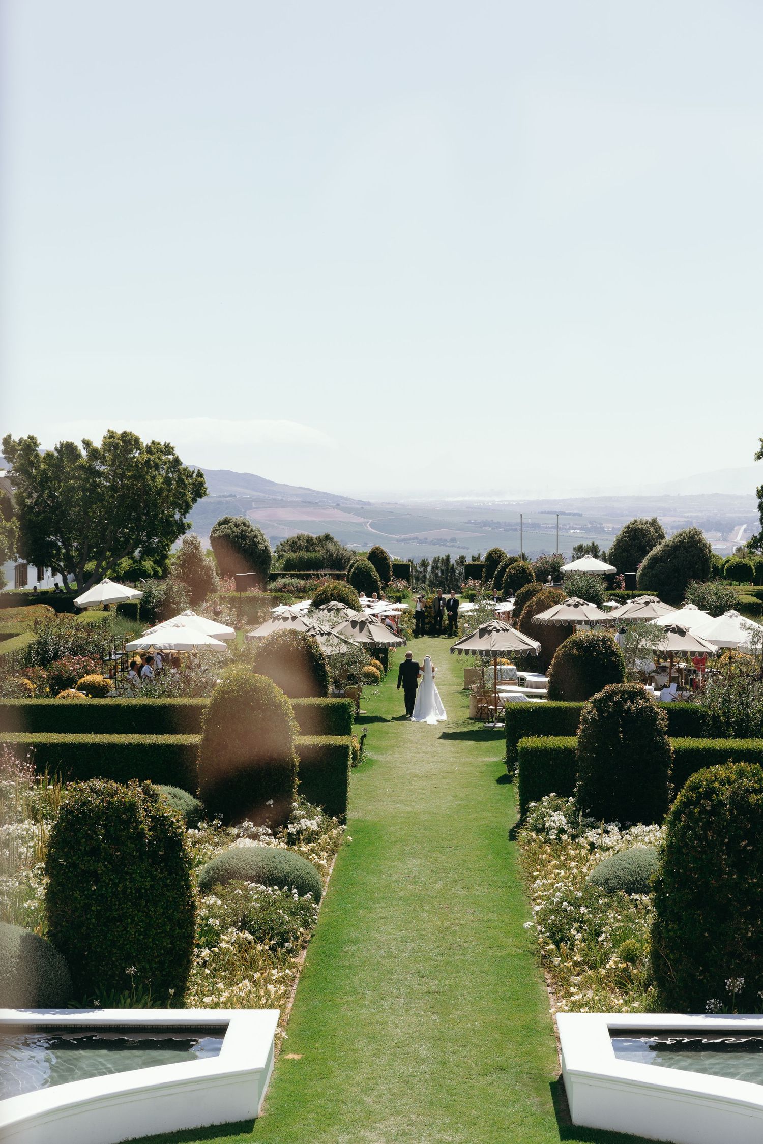 A serene garden pathway with manicured hedges, white benches, and mountain views in the background.