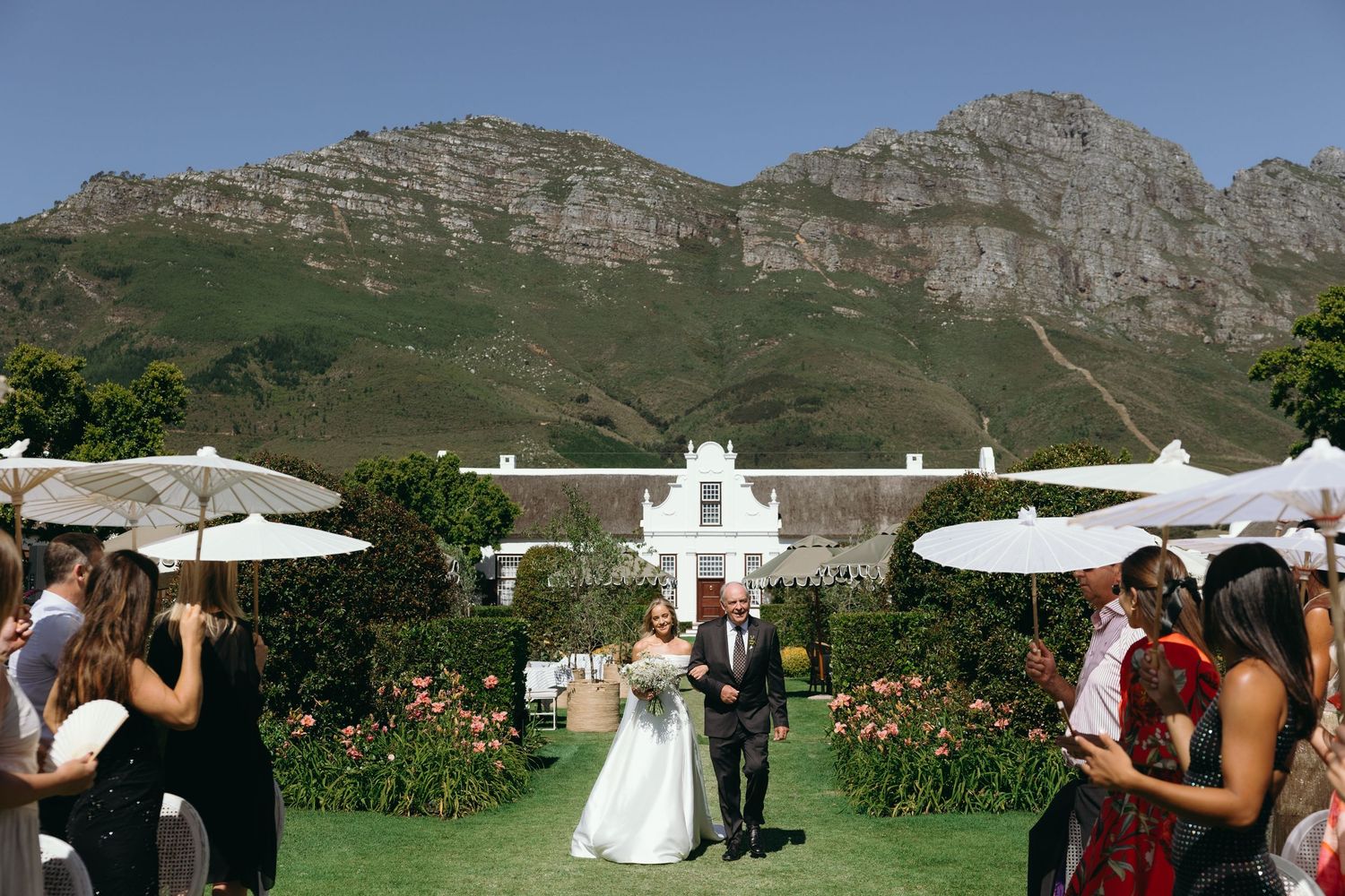 Wedding celebration with white umbrellas and tents set against dramatic mountain backdrop in scenic outdoor venue.