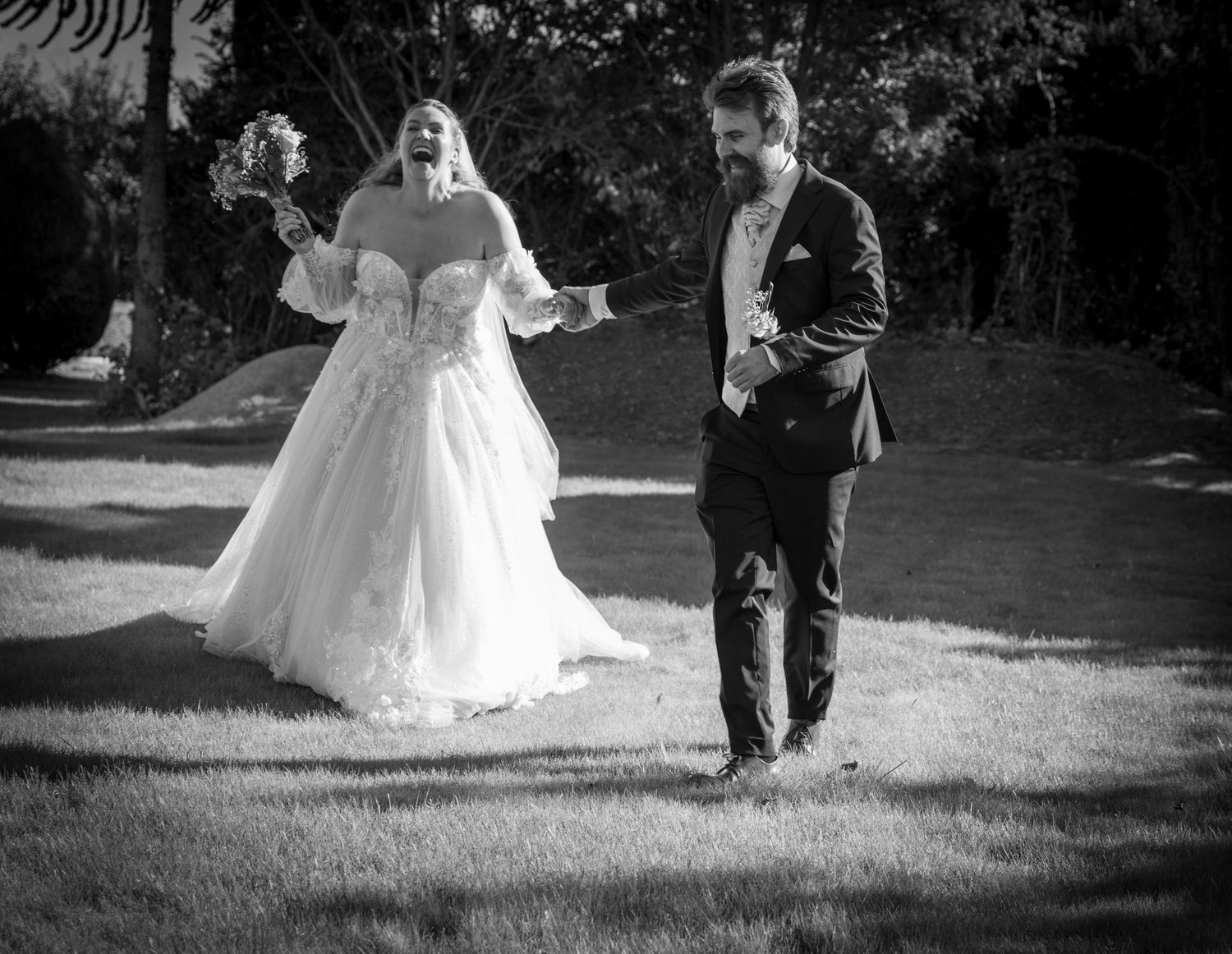 Black and white photo of laughing bride in ball gown approaching groom on a garden lawn.
