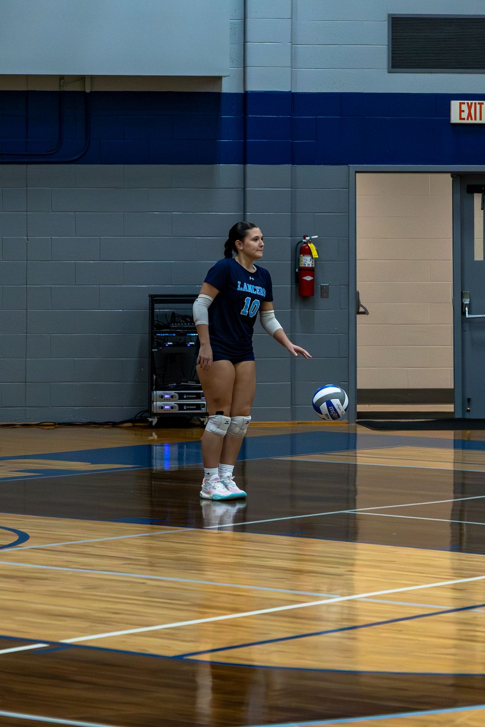 A volleyball player in a gym performs a serve during practice on a wooden court.