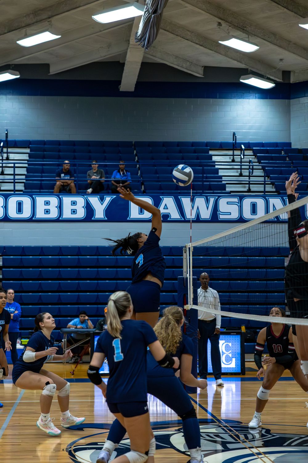 Volleyball team practicing defensive drills and setting techniques on an indoor court with bleachers visible.