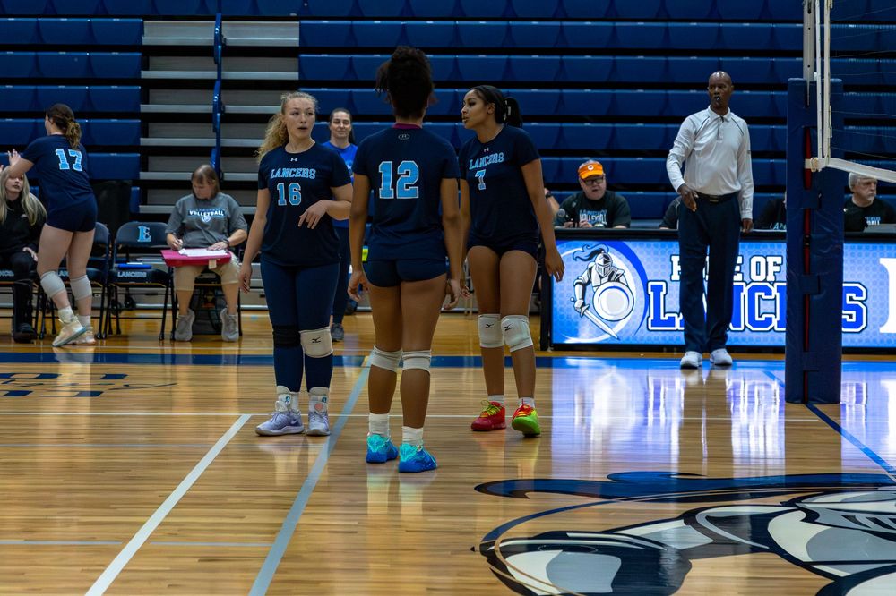 Volleyball players huddle on the court during a game with a scoreboard visible in the background.