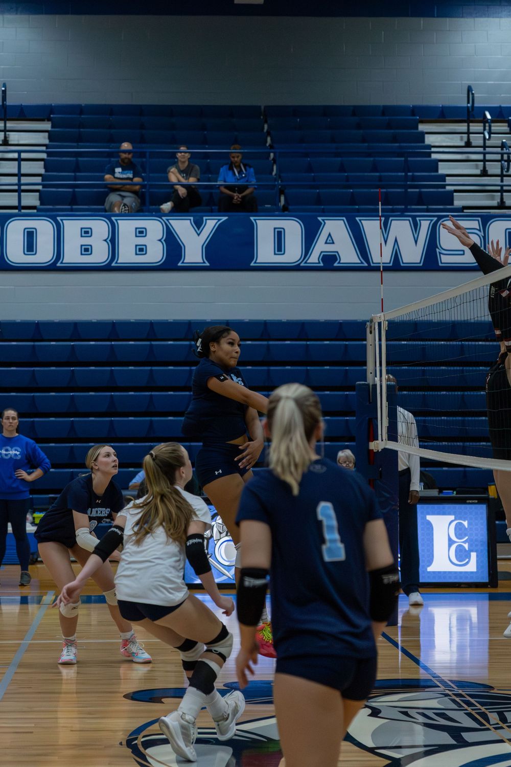 Volleyball players dive and stretch for the ball during an intense indoor match at a blue-themed gymnasium.