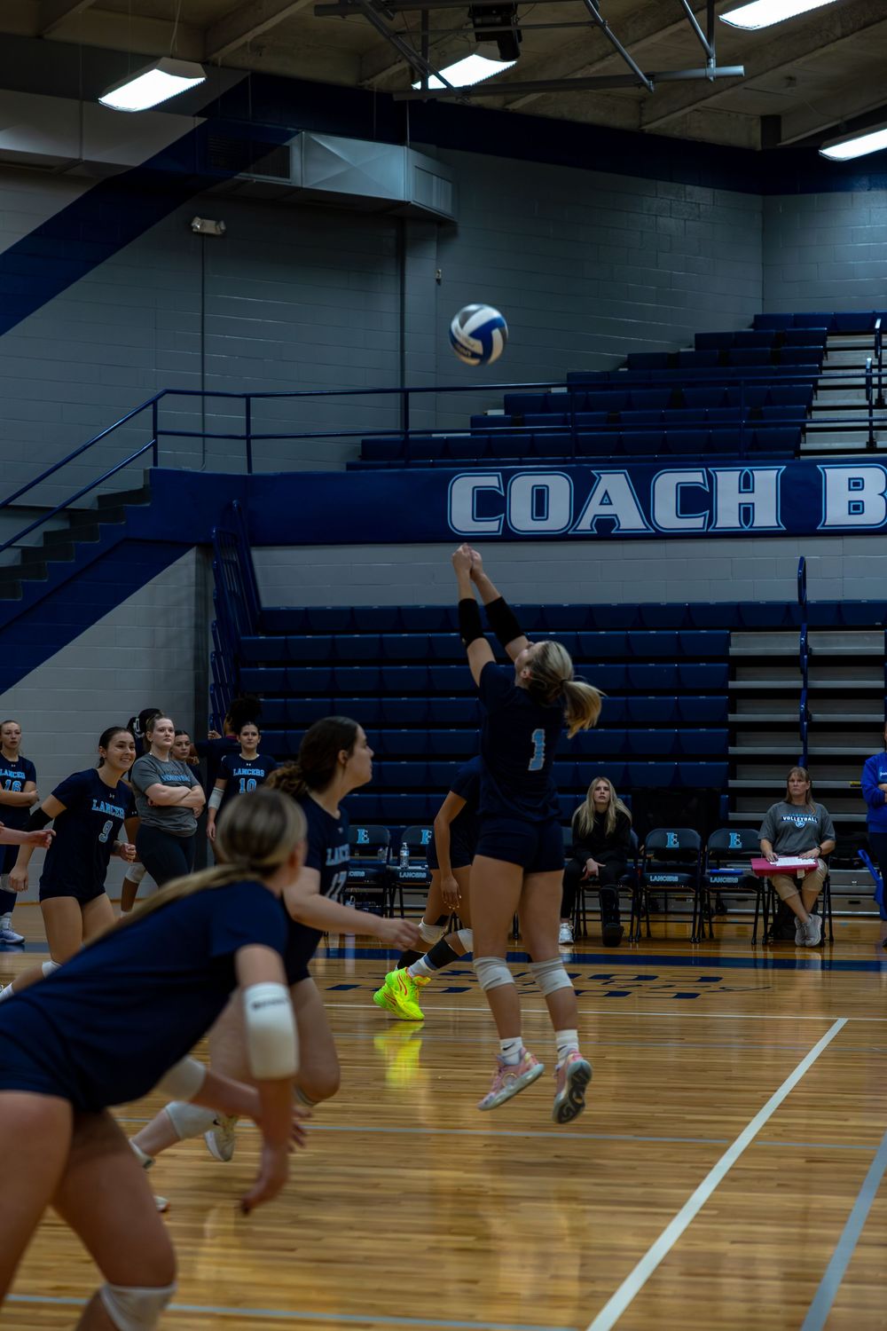 Indoor volleyball players in navy uniforms practicing diving drills on a wooden gymnasium floor.