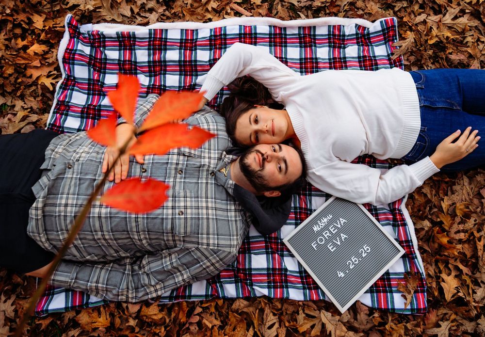 A person lies on a plaid blanket surrounded by autumn leaves while holding a chalkboard sign.