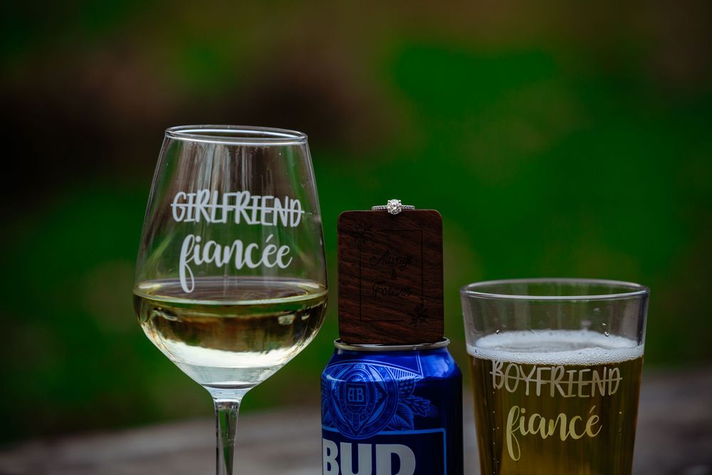 Wine glasses with custom labels for girlfriend and fiance sit on a dark surface with blurred green background.