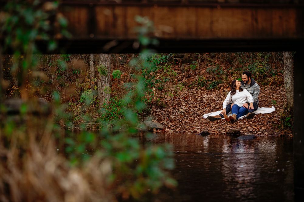 Couple sits together on fallen log by tranquil pond during romantic autumn engagement photo session.