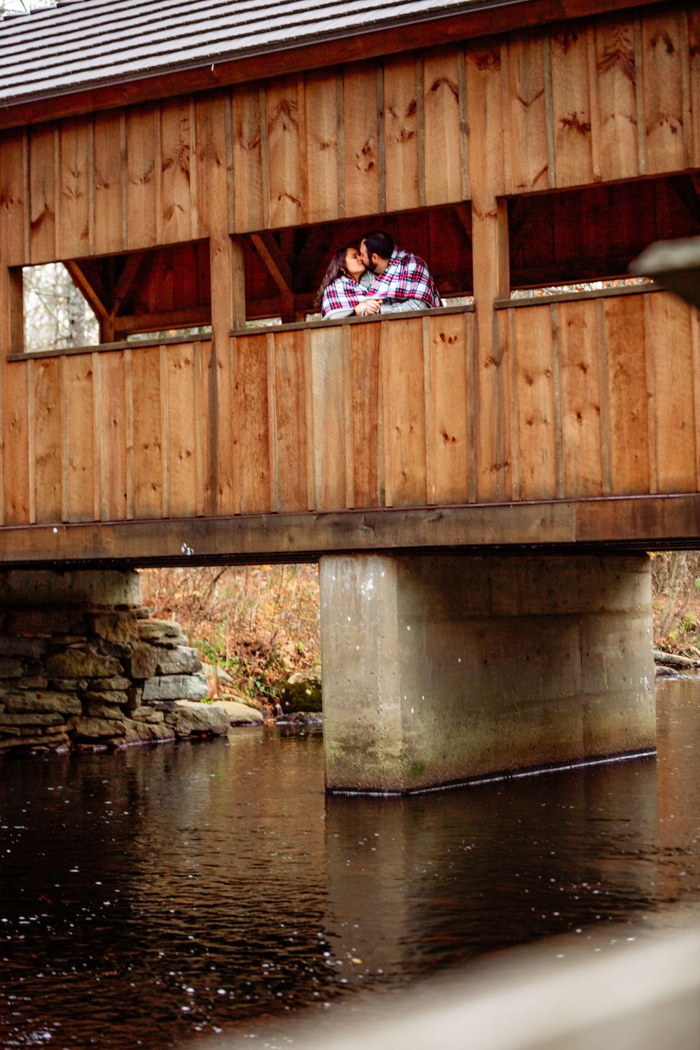 Rustic wooden covered bridge spans across a peaceful river with autumn foliage reflecting in the calm waters.