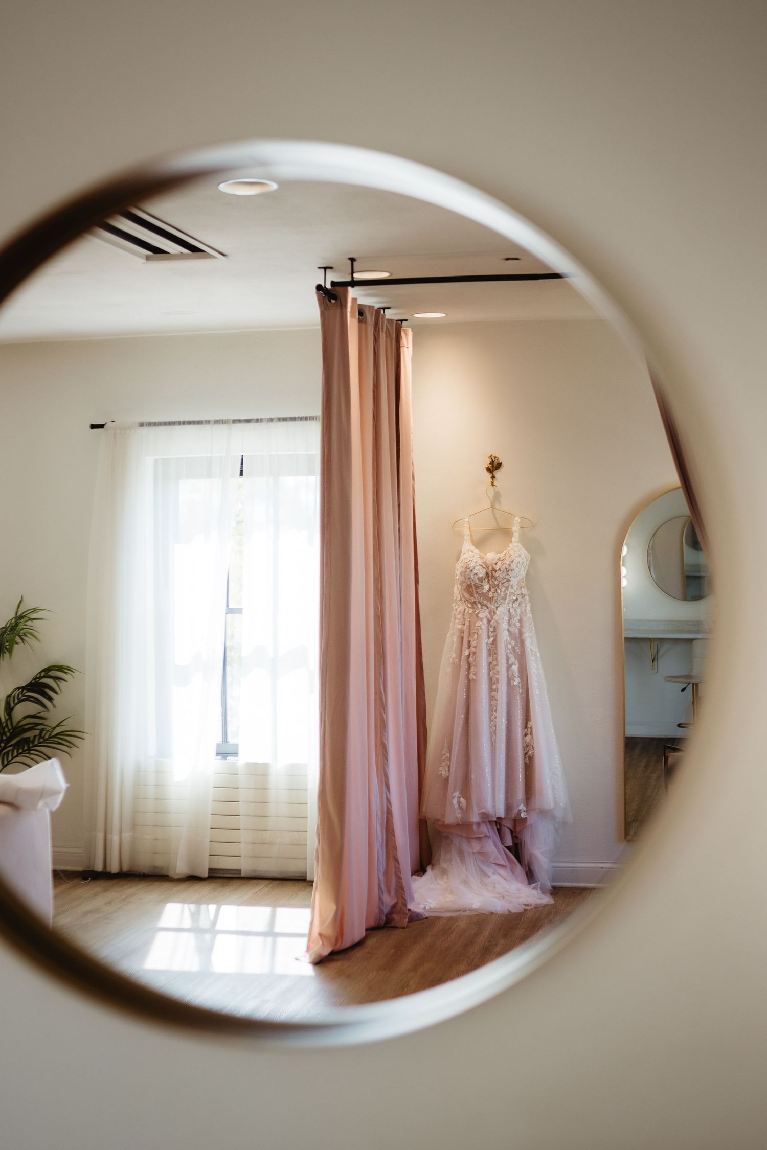 Wedding dress hanging in bridal suite shown through multiple oval mirror reflections with soft pink curtains.