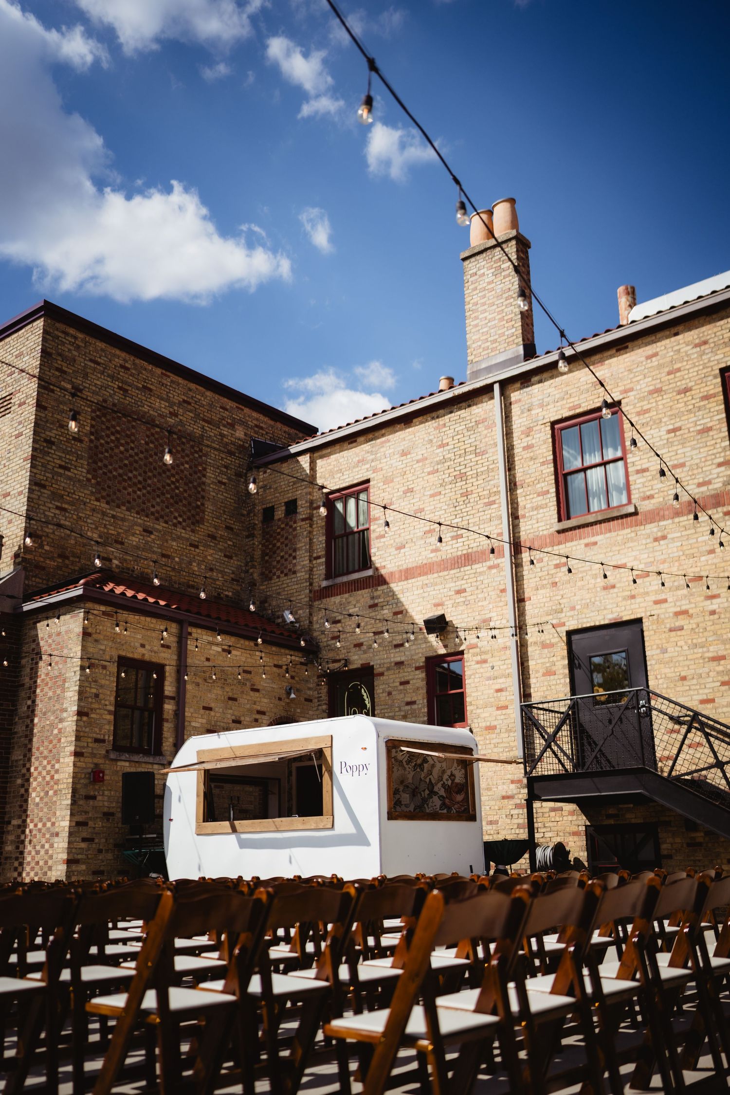 Outdoor wedding venue with rustic brick buildings, rows of wooden chairs, and string lights under a bright blue sky.