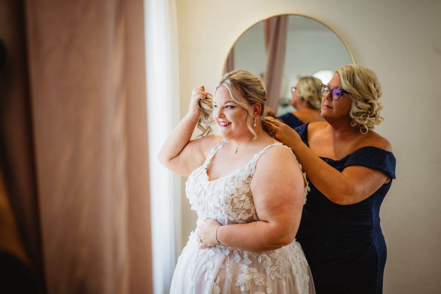 Bridesmaids in navy dresses helping with final wedding preparations in sunlit room.