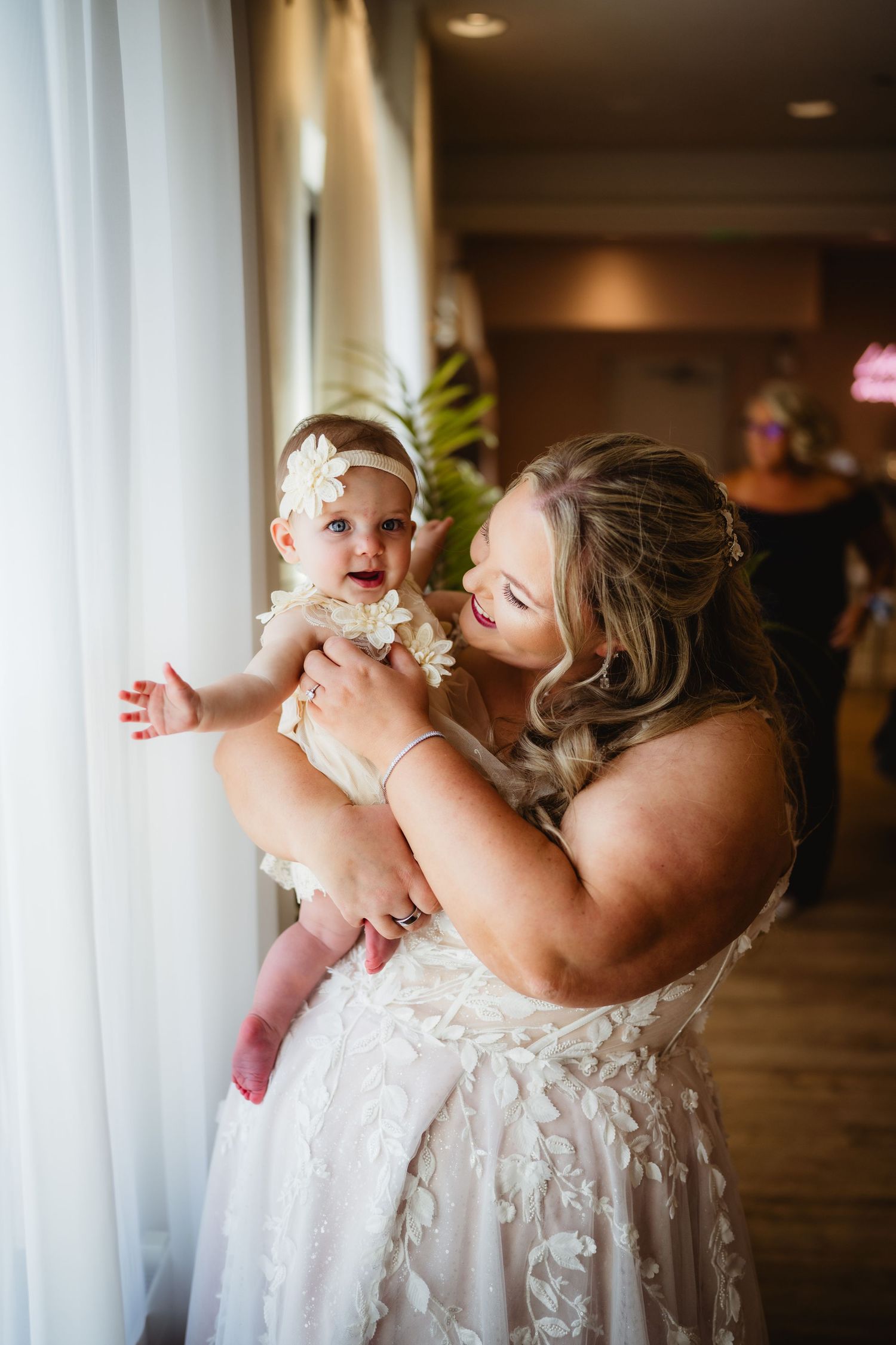 A tender moment shared between a mother and baby in white lace dress against a window backdrop.