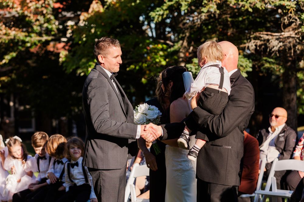 A group of people gathered outdoors for a wedding ceremony, with trees and natural lighting in the background.