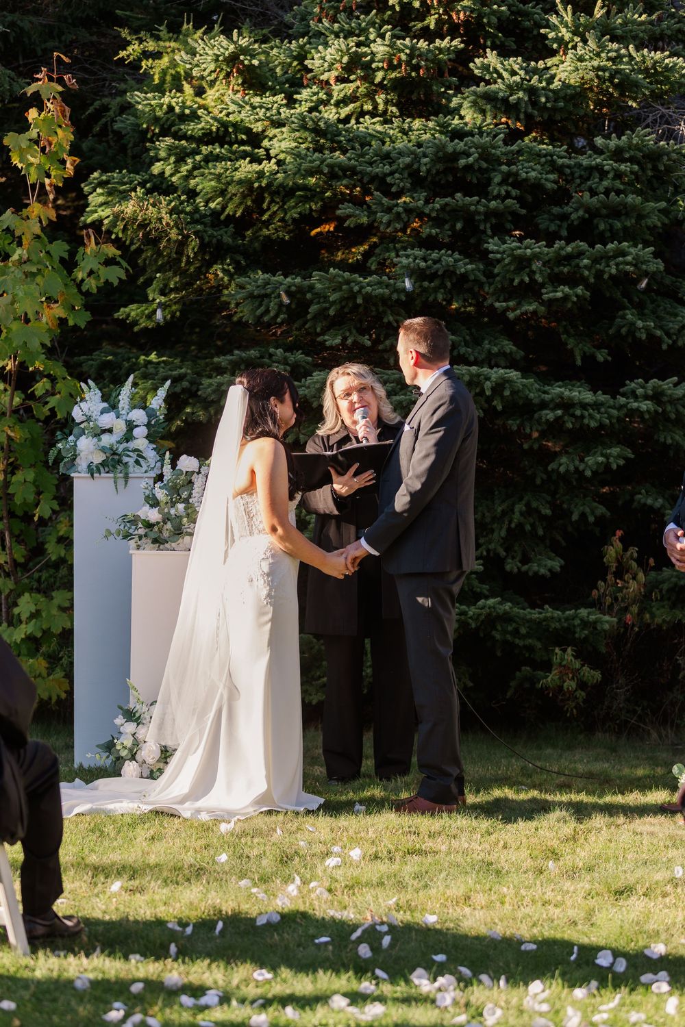 An outdoor wedding ceremony takes place in a garden setting with white floral decorations and greenery in the background.