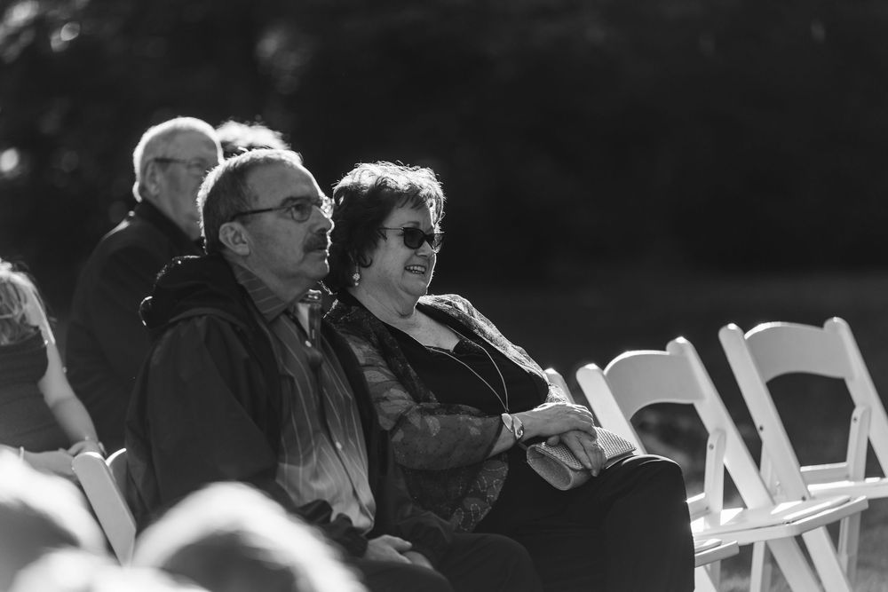 Black and white photo of people seated at an outdoor event with white folding chairs in sunlight.