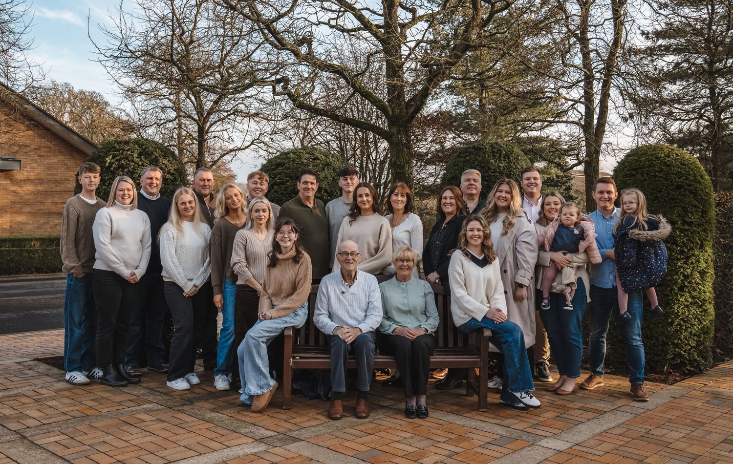 Large extended family group portrait outdoors on a brick patio surrounded by bare winter trees.