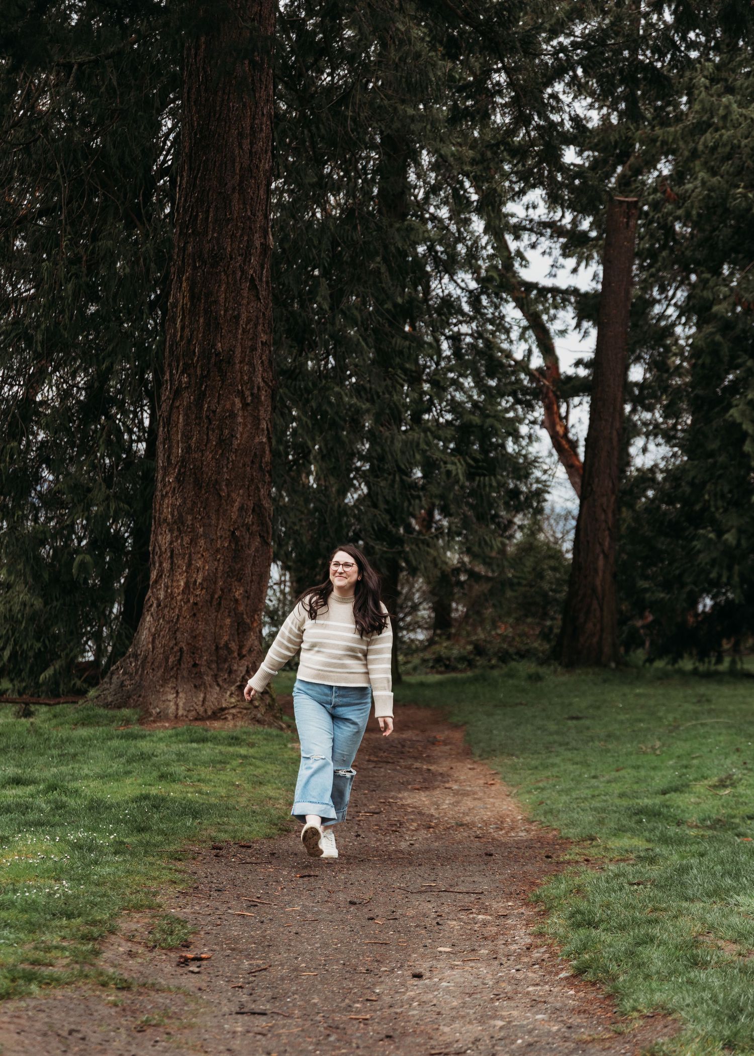 A person walks along a dirt path through a grove of tall evergreen trees on a cloudy day.