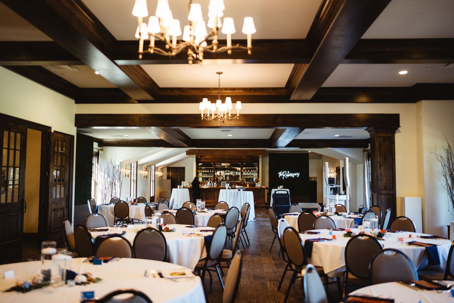 A rustic dining room with exposed wooden beams, chandeliers, and multiple tables set for an elegant gathering.