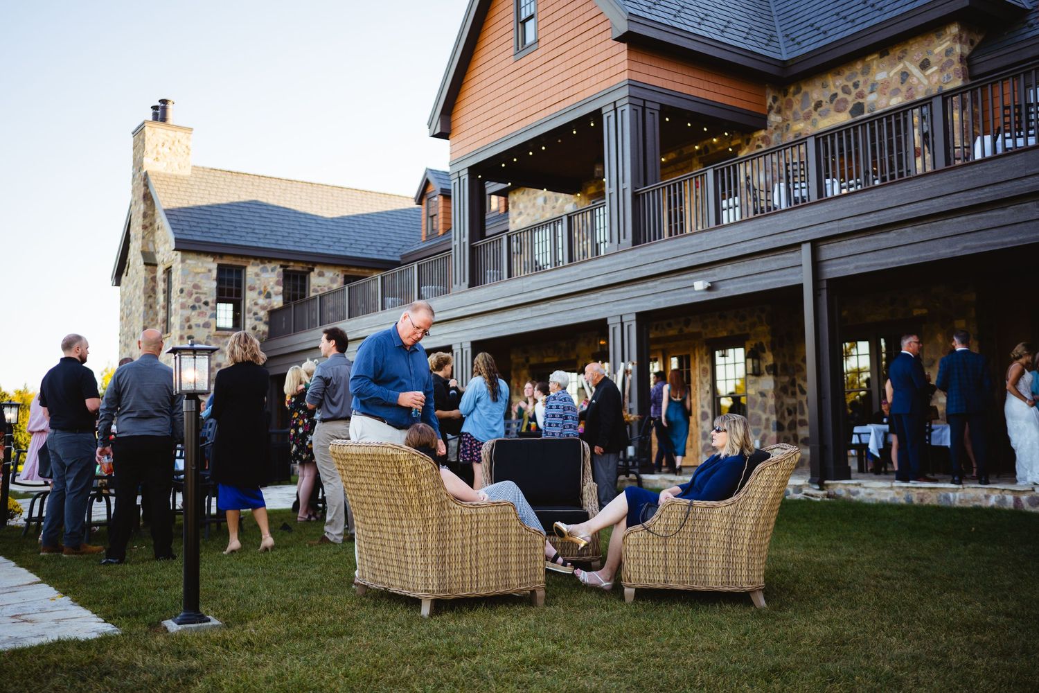 Guests gather on a lawn outside a rustic two-story lodge with stone accents and wraparound deck at dusk.
