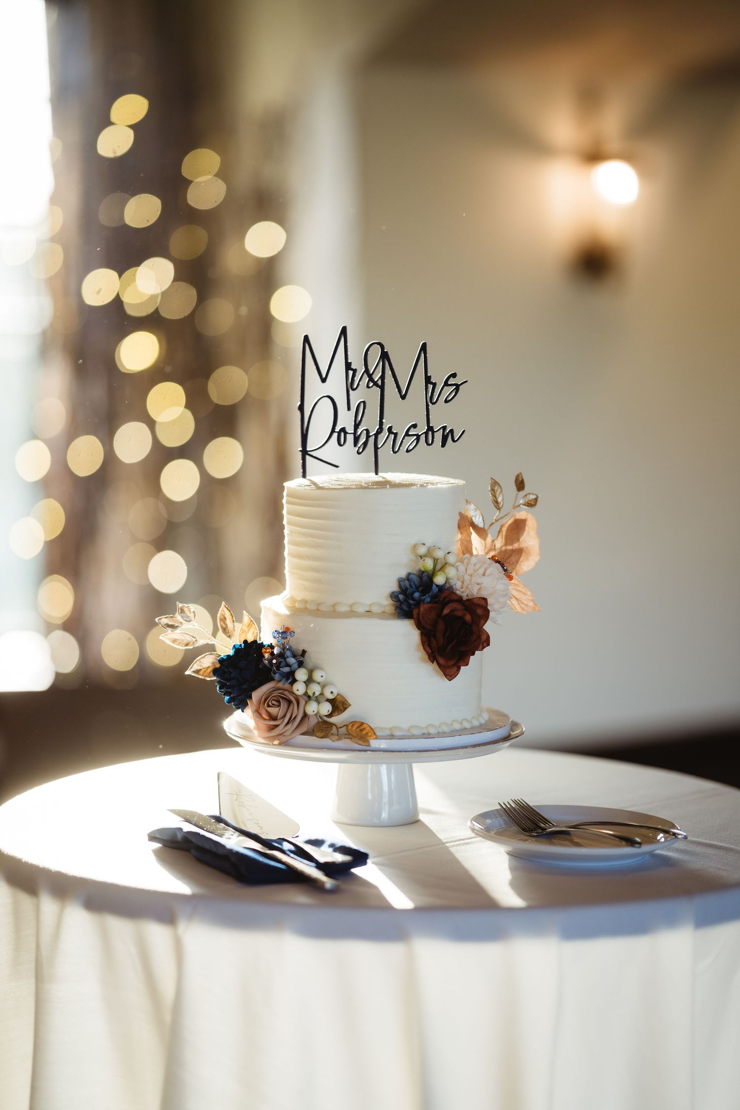 Elegant two-tier white wedding cake with Mr & Mrs Johnson topper and floral accents displayed on white tablecloth.