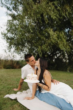 Romantic couple laying on white blanket in grass under tree having intimate moment in outdoor setting.