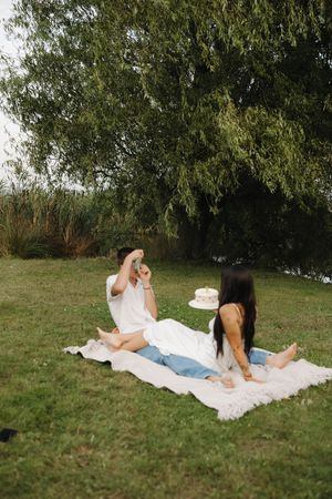 Couple relaxing together on white blanket in grassy outdoor setting during summer day.