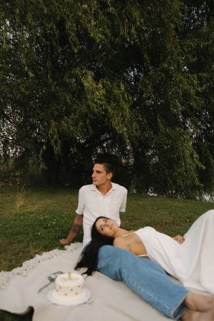 Intimate outdoor photoshoot of couple laying on white blanket under tree branches.