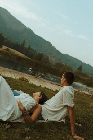 Couple in white shirts laying on grass with mountain backdrop during romantic photoshoot.