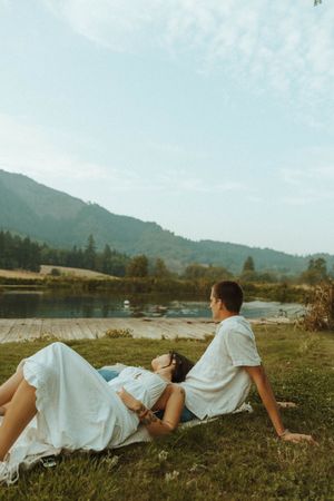 Romantic couple pose on grassy hillside with mountains and lake in background.