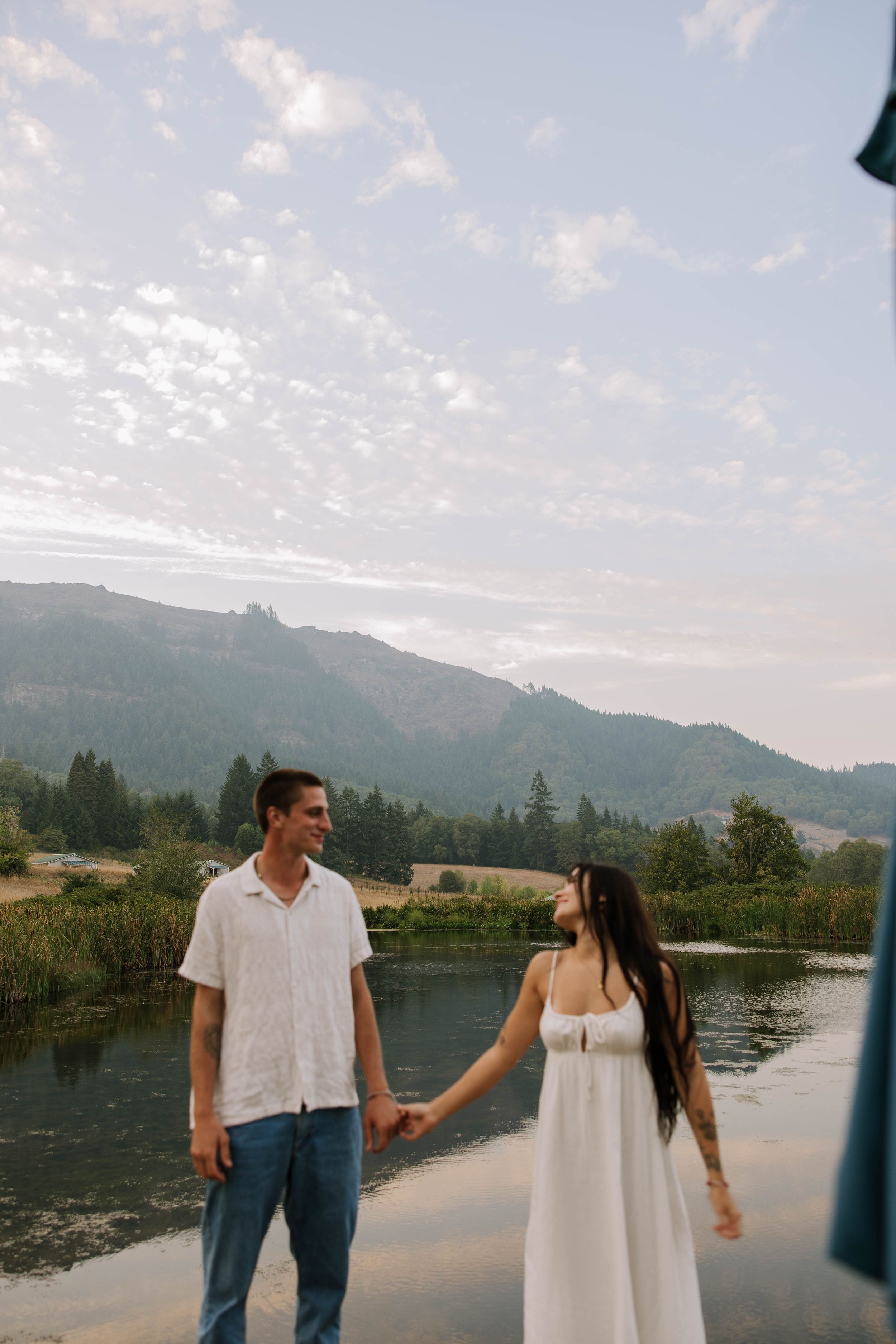 Couple holds hands on dock with mountain landscape and lake in background.