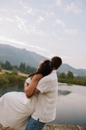 Couple embraces on lakeside dock with mountain scenery at dusk.