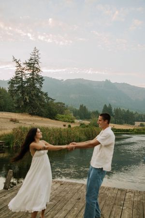 A couple dances playfully on a wooden dock by a lake with mountains in the background at sunset.