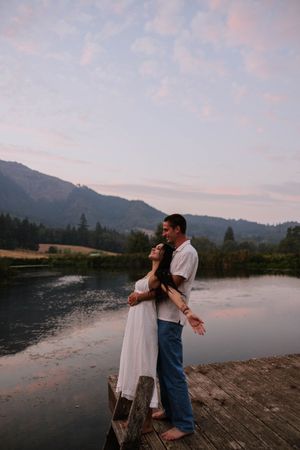 A couple embraces on a wooden dock with mountains and a lake in the background during sunset.