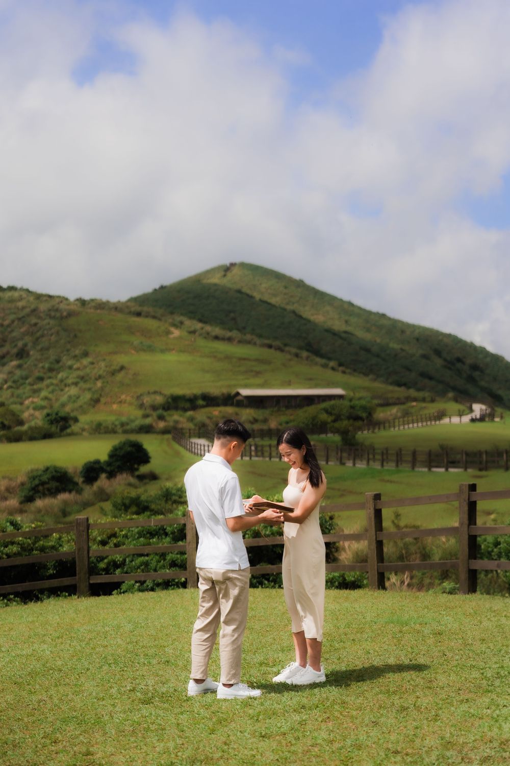 A couple in white and beige outfits share a romantic moment in a scenic green grassland at Taiwan's Qingtiangang in Yangminshan Park.
