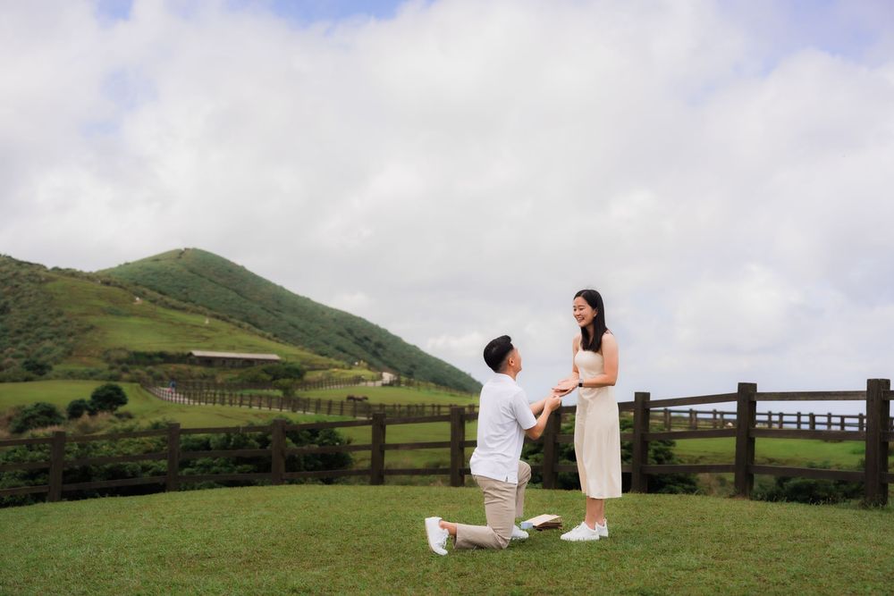 A couple in light colored attire in mid marriage proposal with scenic green rolling hills on a beautiful sunny day.