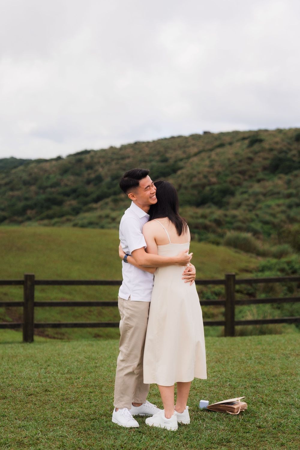 A couple shares a tender embrace while standing in a green meadow with rolling hills in the background.