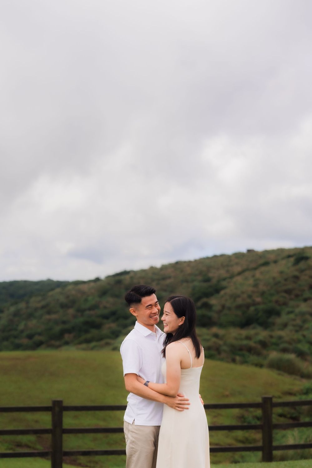 A couple shares a romantic moment against a scenic green hillside backdrop with wooden fence at Qingtaingang Grassland in north Taiwan.