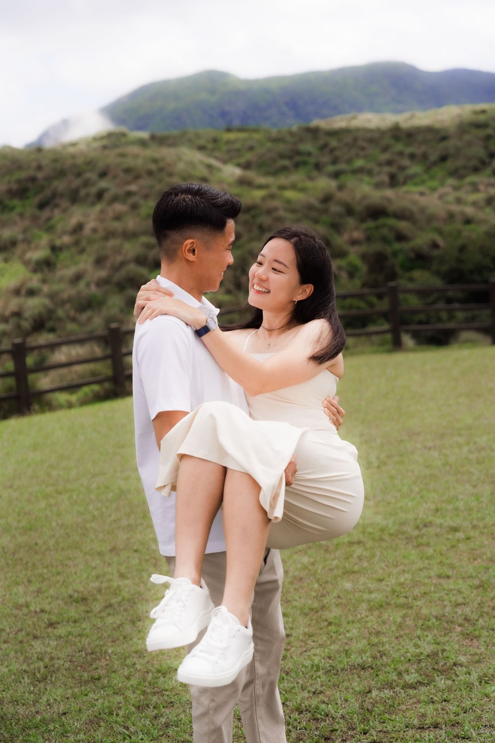 A couple in coordinated white outfits embrace playfully in a grassy field with mountains in the background.