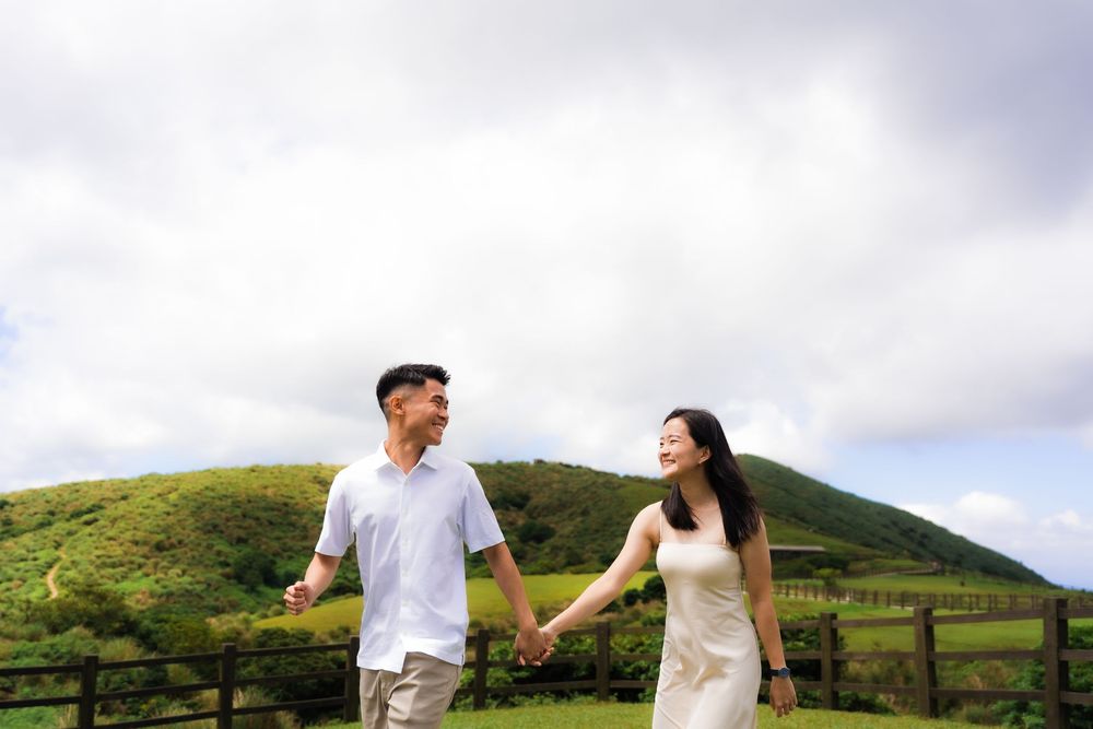 A couple in coordinate white attire walks hand in hand through lush green hills on a white cloudy day in Taiwan's Yangmingshan National Park