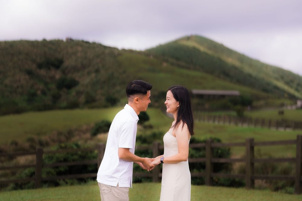 A couple just engaged holds hands in excitement standing in a field against rolling green hills and scenic landscapes.