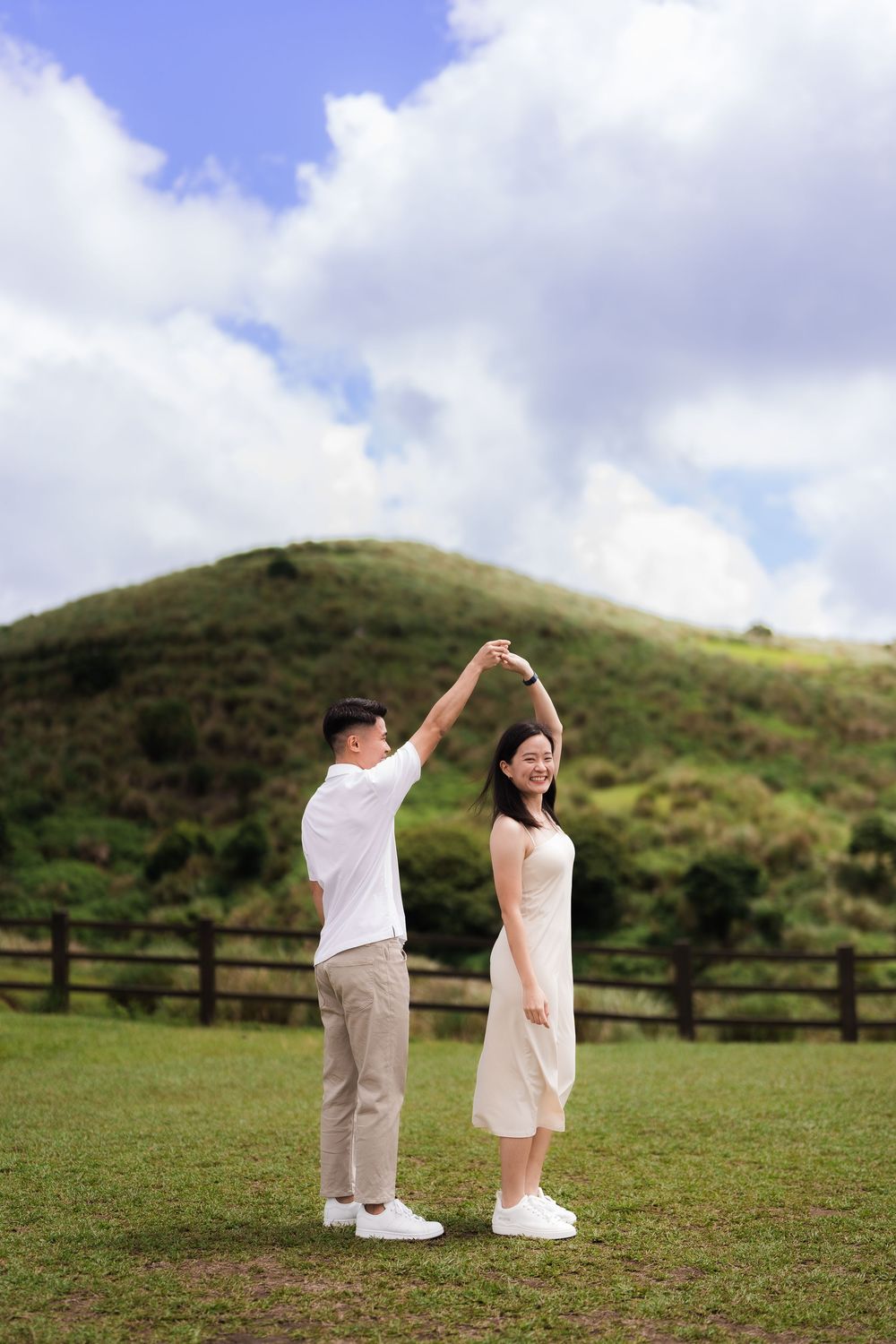 A man twirls his new fiancé in a beautiful open grass field with rolling green hills and a cloudy blue sky in the background.