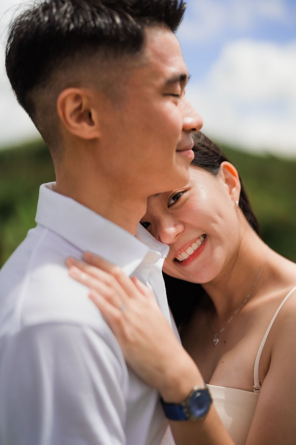 A young woman rests her head against a young man's shoulder and gently lays her hand showing a beautiful engagement ring on his chest.