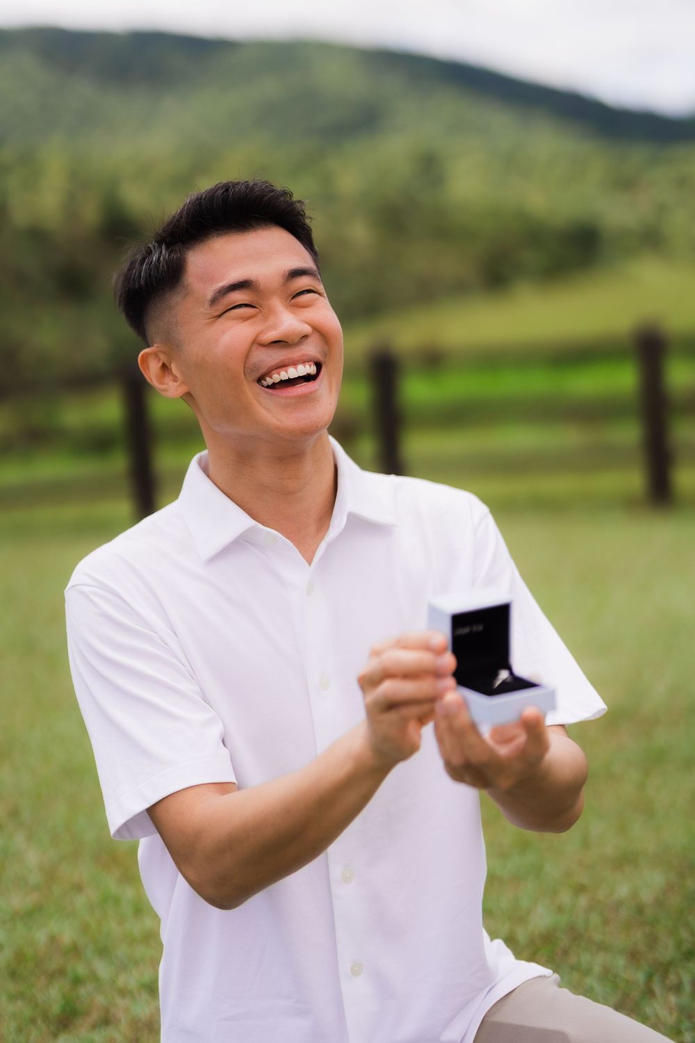 A joyful man wearing light colors kneels down on a beautiful grassy meadow holding an open ring box about to propose.