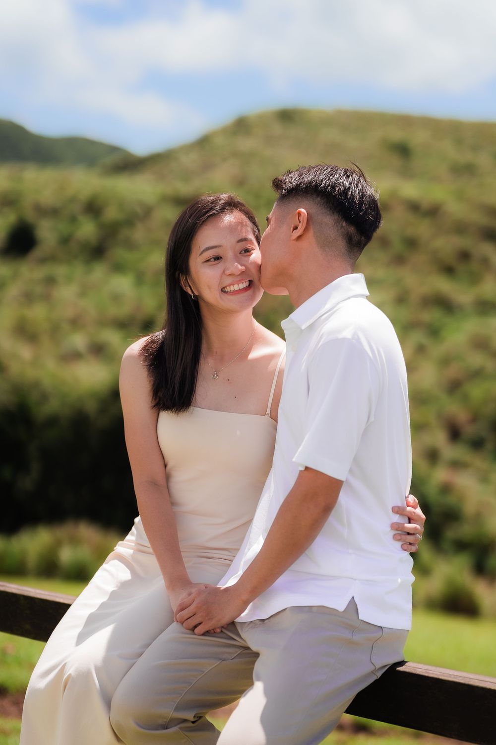 A couple just engaged sneak a kiss while sitting on a rustic wooden fence against luscious rolling green hills in the background.