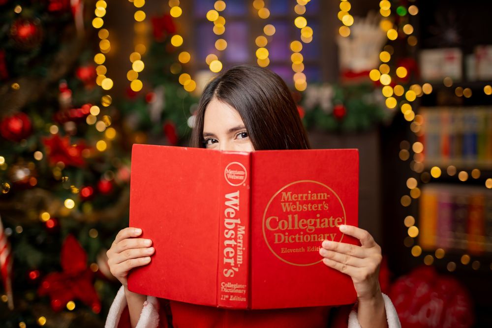 A person in festive red attire holds a red book while sitting in front of Christmas decorations and twinkling lights.