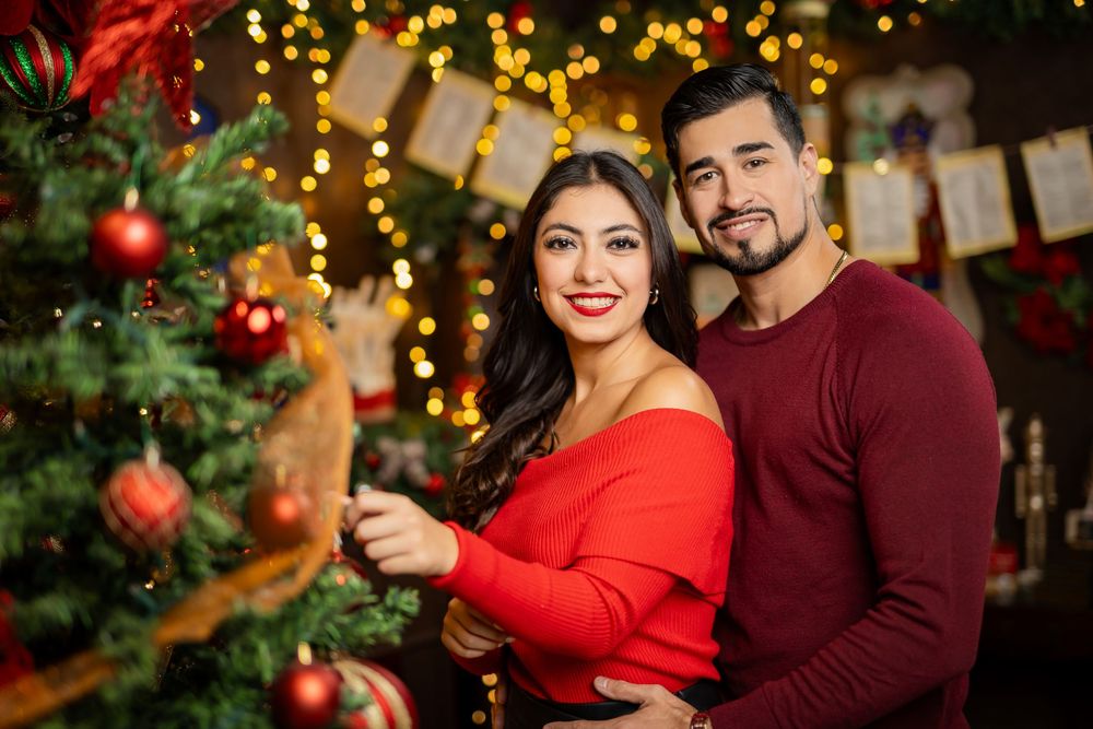 A couple in festive red and burgundy attire poses near a decorated Christmas tree with twinkling lights in the background.